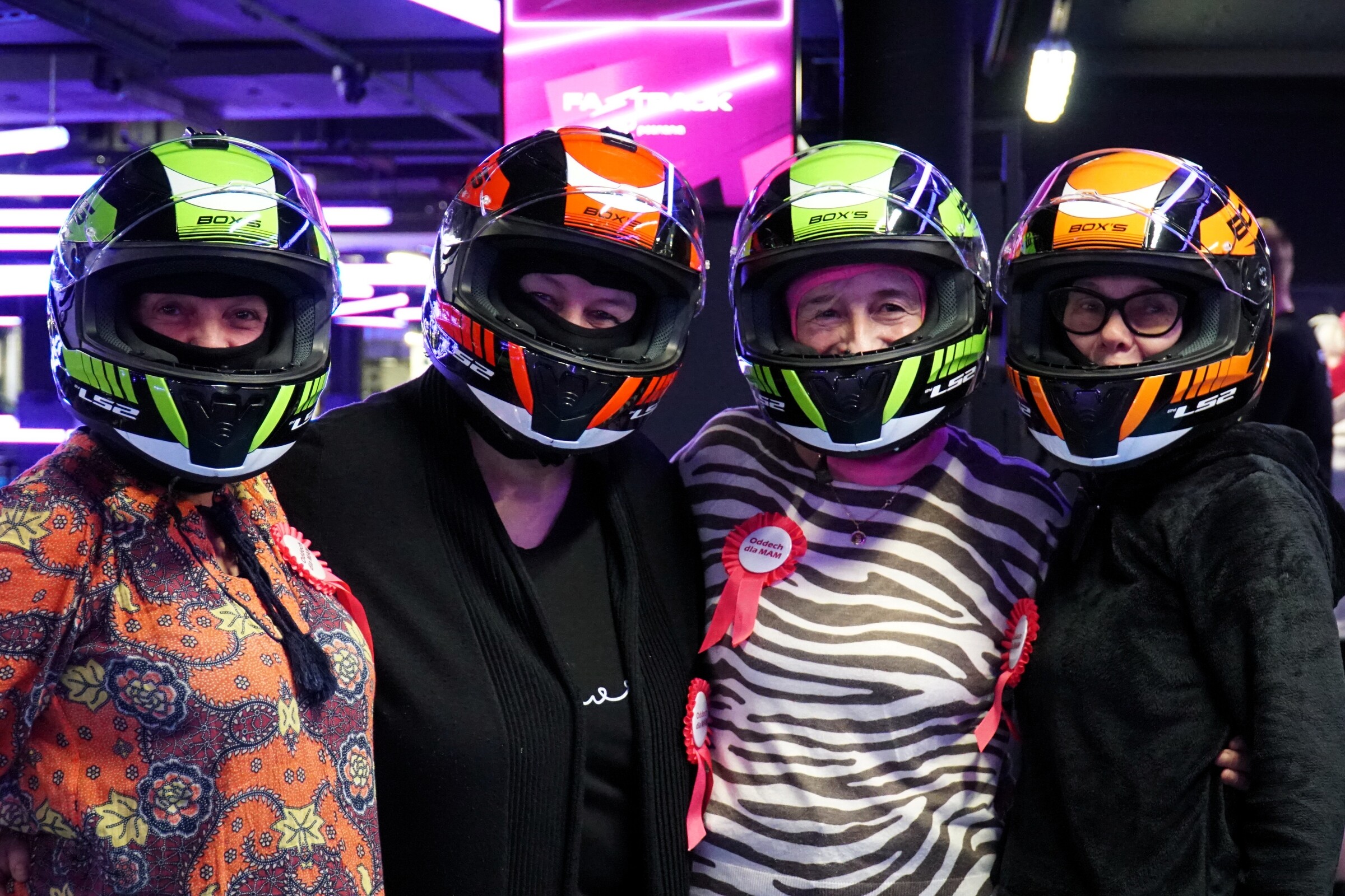 Cztery kobiety w kaskach ochronnych stoją obok siebie, uśmiechając się do zdjęcia. W tle tor gokartowy Fast Track i kolorowe światło.